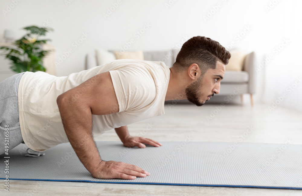 Fototapeta premium Side view of young athletic Arab man doing push-ups on sports mat at home, copy space