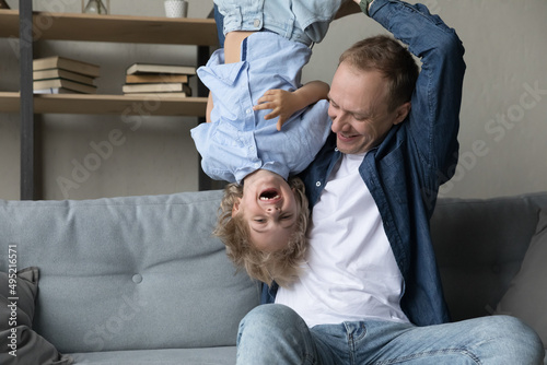 Happy young father lifting in air upside down laughing adorable small kid son, having fun together in living room. Joyful two male generations family enjoying entertaining activity on weekend at home.