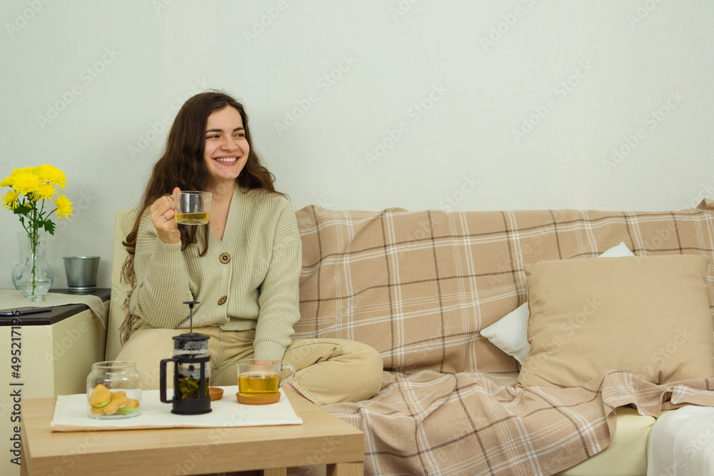 In a room with yellow flowers in a vase, a young woman sits on a sofa at a tea table with a mug of tea in her hand, smiles broadly and cheerfully, looks away in a good playful mood, copy space.