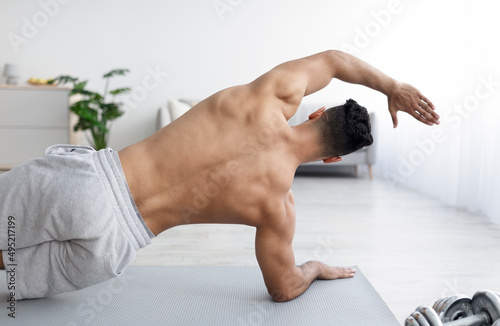 Back view of young Arab guy with bare torso standing in side plank on yoga mat, training muscles at home