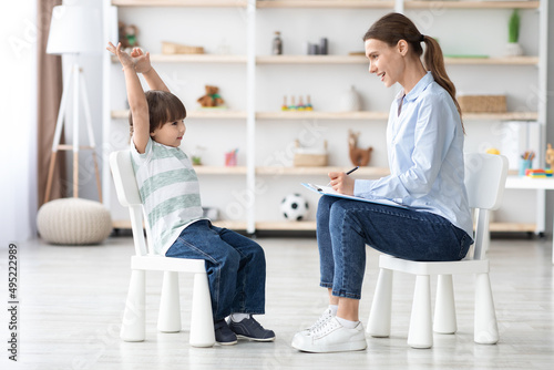 Cute talkative little boy telling story to professional woman psychologist during consultation, therapist taking notes photo