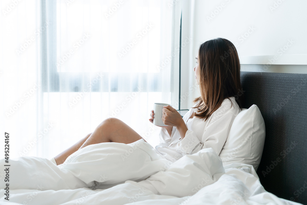 Young beautiful brunette hair female in white shirt pajamas drinking coffee while sitting on the bed in the morning. Young beautiful brunette hair female in white shirt pajamas drinking coffee while sitting on the bed in the morning.