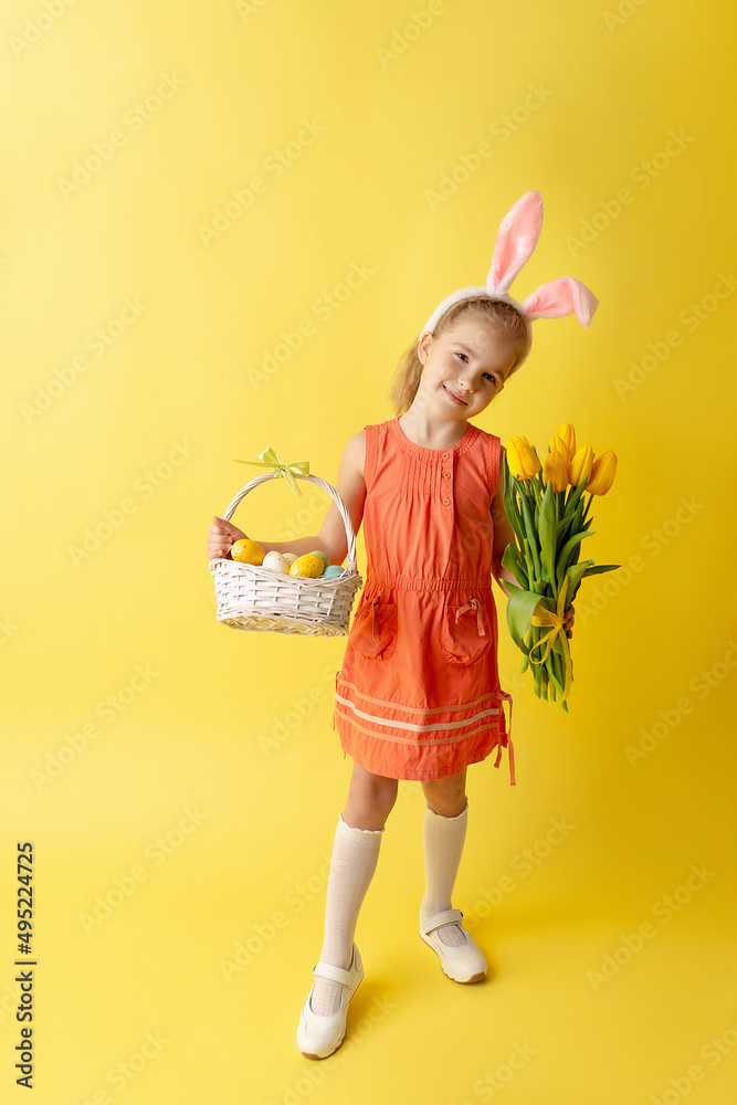 Beautiful cute little girl in Easter bunny ears holds a basket with eggs and spring bouquet on a yellow background Beautiful cute little girl in Easter bunny ears holds a basket with eggs and spring bouquet on a yellow background