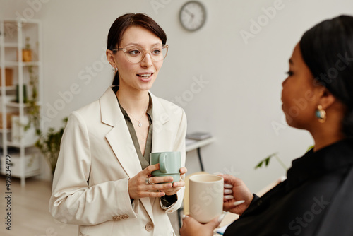 Minimal portrait of two successful young women talking at coffee break in office, focus on Caucasian businesswoman wearing glasses