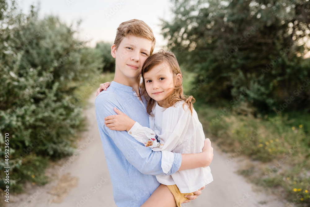 Fototapeta premium Portrait of older brother and younger sister together outside. Siblings enjoying summertime outside in the field. Natural facial expression, minimal editing photo