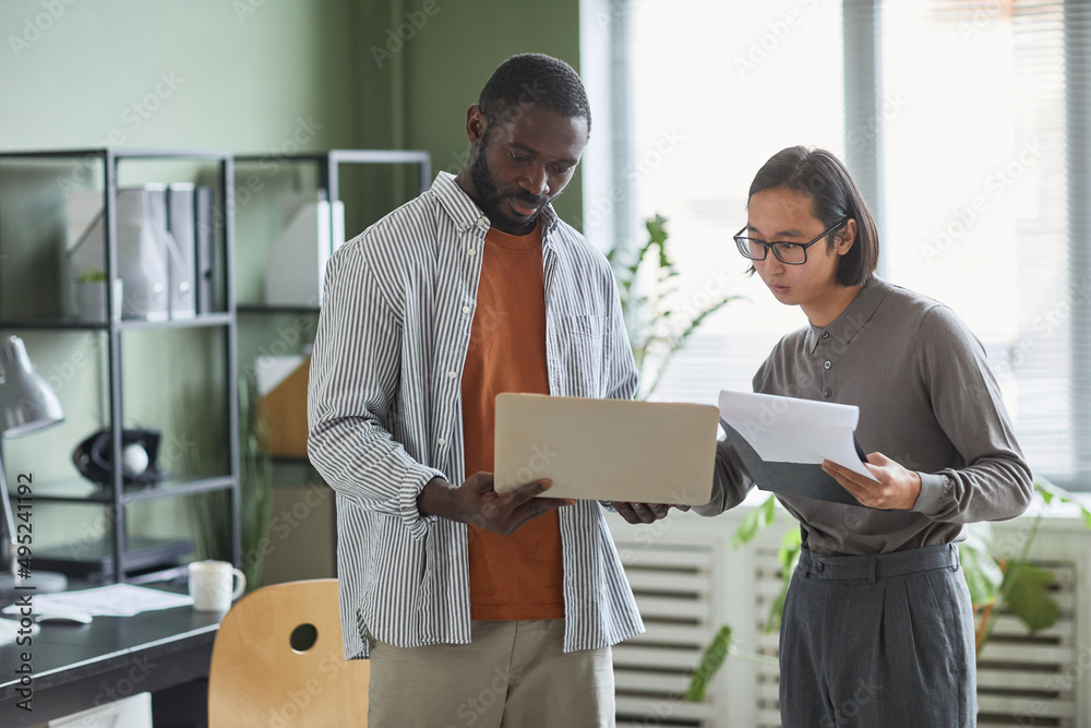 Fototapeta premium Waist up portrait of two ethnic business people African American and Asian discussing project while standing in modern office