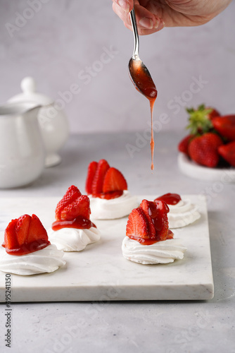 Pouring red jam over mini-pavlova with fresh strawberries - delicious meringue cakes on marble board on off-white background