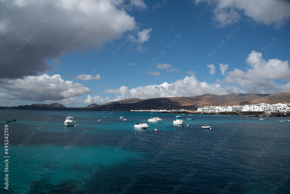 Hermoso mar azul turquesa y transparente con algunas barcas y casas blancas tradicionales en la parte de atrás con un cielo azul y montañas volcánicas en la parte de atrás en Lanzarote, Islas Canarias