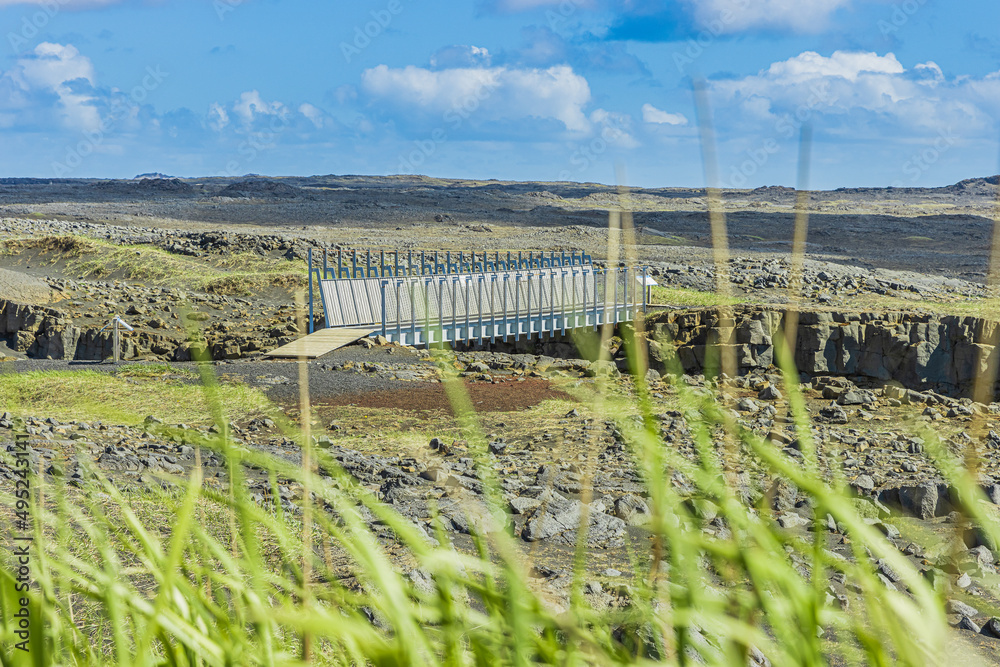 Metal bridge over the American and Eurasian tectonic plates in sunshine. Volcano landscape on the Reykjanes Peninsula of Iceland. View through green blades of grass in the foreground