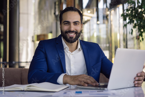Handsome smiling Moroccan businessman using laptop computer working online sitting in modern office. Portrait of happy successful middle eastern manager looking at camera sitting at workplace 