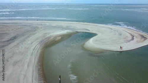 Aerial view over gentle surf, flying over a curved spit of land as beachgoers walk on the sand.