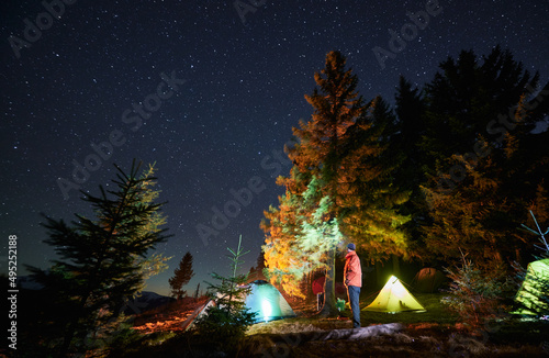 Caucasian male wanderer admire beauty of mountains during night time with starry sky, active hiker man in professional wear recreating in campground during mountaineering vacations.