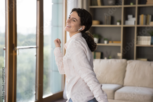 Happy joyful active Latin girl with wireless earphone in ear enjoying favorite songs, tunes, dancing in living room, listening to music with online app, service, smiling. Cheerful woman at home party
