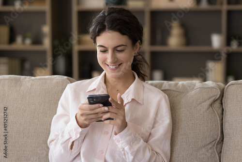 Pretty young millennial customer girl using smartphone on home couch, relaxing on sofa in living room, buying online, shopping on internet virtual ecommerce app on mobile phone, texting message