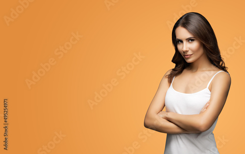 Portrait of woman in white casual tank top wear, standing in cross arms pose, over vivid brown background. Brunette girl at studio. Wide composition with copy space free area for some text.