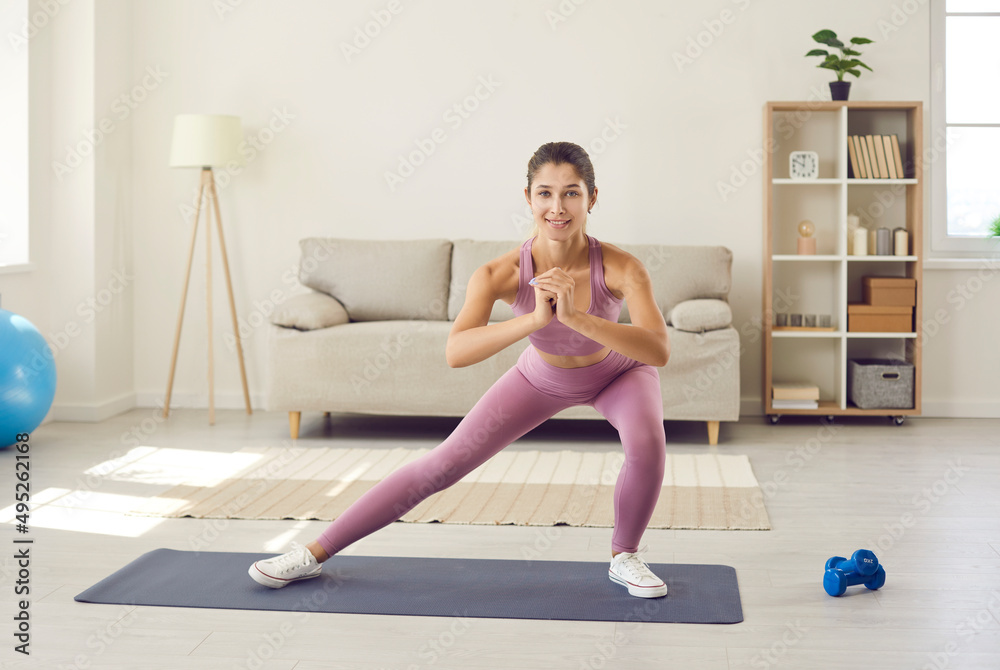 © Studio Romantic - Happy beautiful slim woman enjoying routine fitness workout at home. Active young Caucasian lady in modern sports leggings standing on gym mat in living room, smiling and doing side lunge exercise