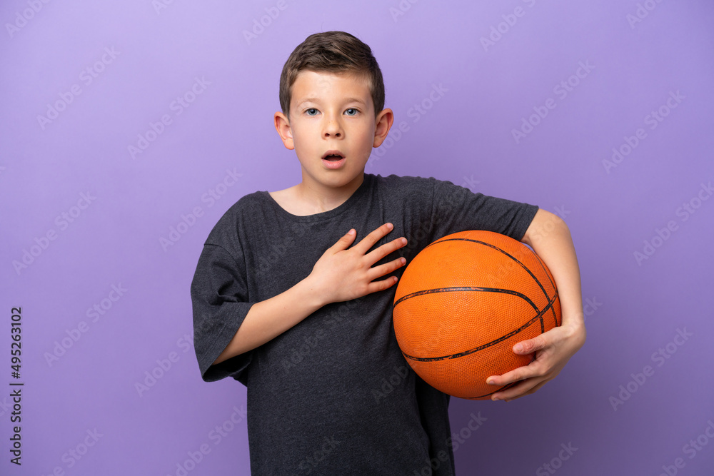 Little boy playing basketball isolated on purple background surprised and shocked while looking right Little boy playing basketball isolated on purple background surprised and shocked while looking right