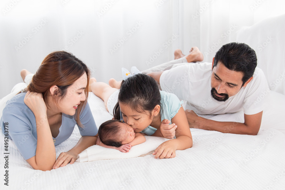 Asian parents and newborn with sister spend time together in bedroom. Sister kiss Adorable infant while baby sleeping among happy family, mother and father lying in bed with two daughters.