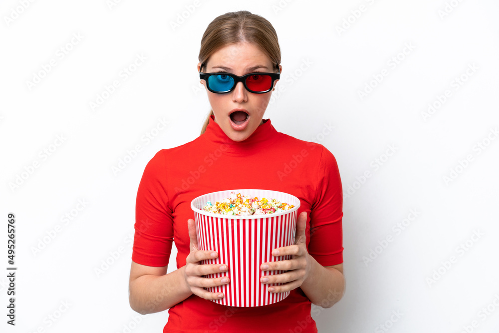 Young caucasian woman isolated on white background surprised with 3d glasses and holding a big bucket of popcorns