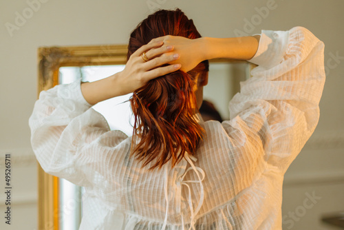 A beautiful red-haired girl in the bathroom looks in the mirror and ties her ponytail. Pretty woman stroking her hair while looking in the mirror. The concept of hairstyles and hair care. Back view