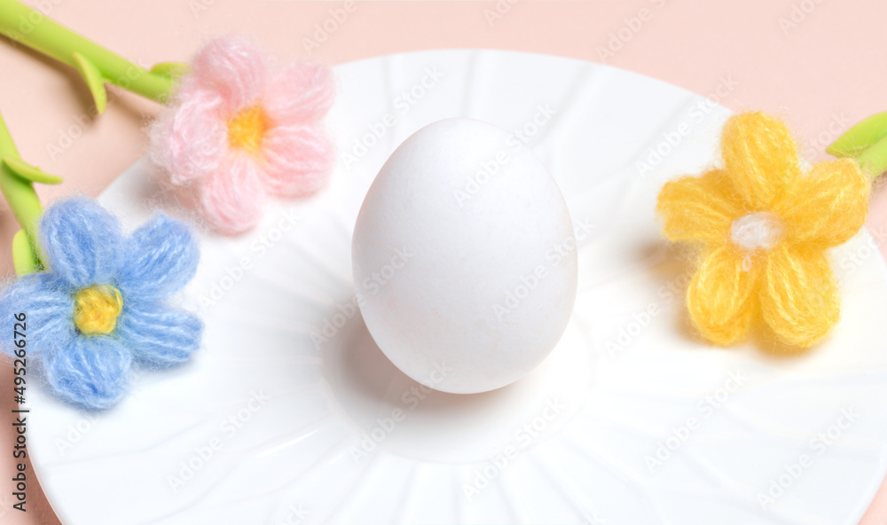 Easter minimal banner with white egg, knitted color flowers on white plate on pastel pink backdrop.
