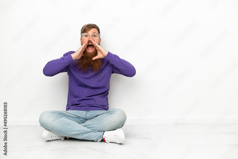 Young caucasian reddish man sitting on the floor isolated on white background shouting and announcing something Young caucasian reddish man sitting on the floor isolated on white background shouting and announcing something