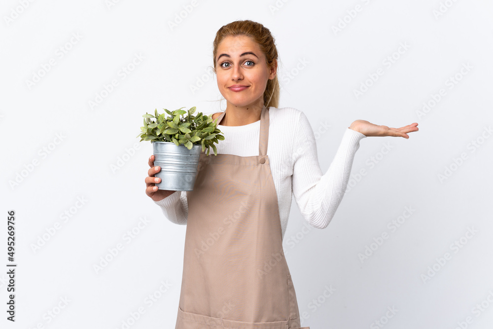 Young blonde gardener woman girl holding a plant over isolated white background having doubts with confuse face expression Young blonde gardener woman girl holding a plant over isolated white background having doubts with confuse face expression