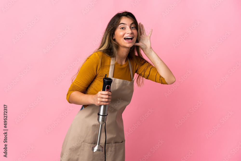Young chef woman using hand blender isolated on pink background listening to something by putting hand on the ear Young chef woman using hand blender isolated on pink background listening to something by putting hand on the ear