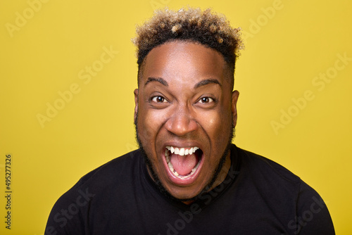 An enraged black guy screams loudly with his mouth wide open and bulging eyes. An outraged African-American yells with all his might in outrage close-ups on camera on a yellow background. © Aleksandr