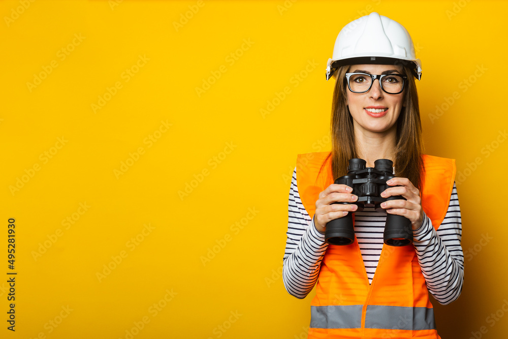 Young woman with a smile in a vest and a helmet holds binoculars on a yellow background. Construction concept, new building. Banner