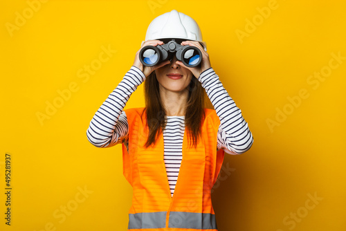 Young woman in a vest and a helmet looks through binoculars on a yellow background. Construction concept, new building. Banner