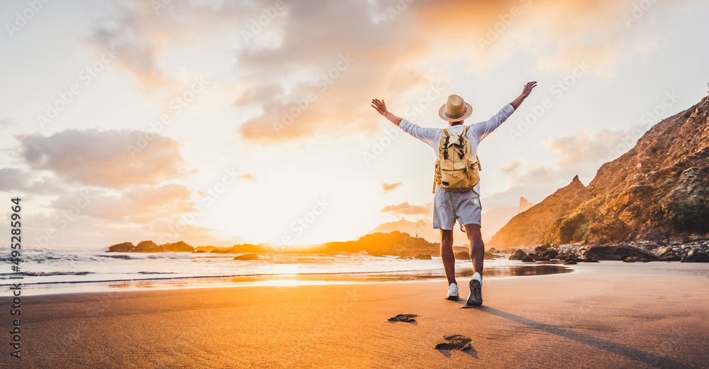 Plakat Happy man with hands up enjoying wellbeing and freedom at the beach - Male with backpack traveling in the nature with sunrise view - Healthy lifestyle, happiness and travel concept