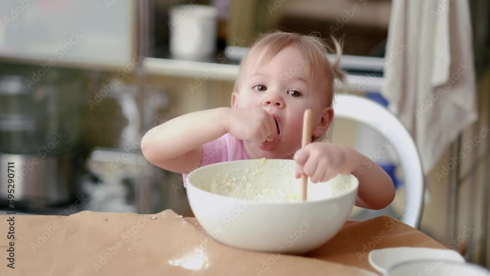 Little cute funny girl licking the dough from her finger helping mother prepare pie cake in kitchen, baking homemade cookie together Little cute funny girl licking the dough from her finger helping mother prepare pie cake in kitchen, baking homemade cookie together