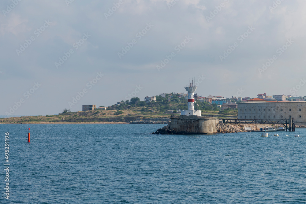 White lighthouse stands on the coast of Black sea. Sunny day. Small waves on the sea water surface. Clouds on the sky. Navigation safety theme.
