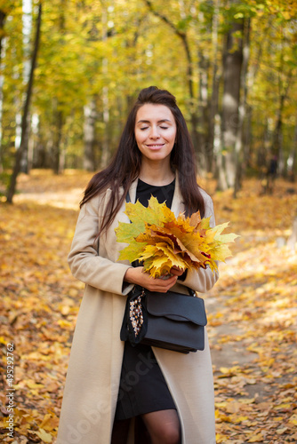 Brunette woman in autumn park and autumn leaves. Small black bag. Autumn mood. Yellow  red and green leaves. Beige coat.