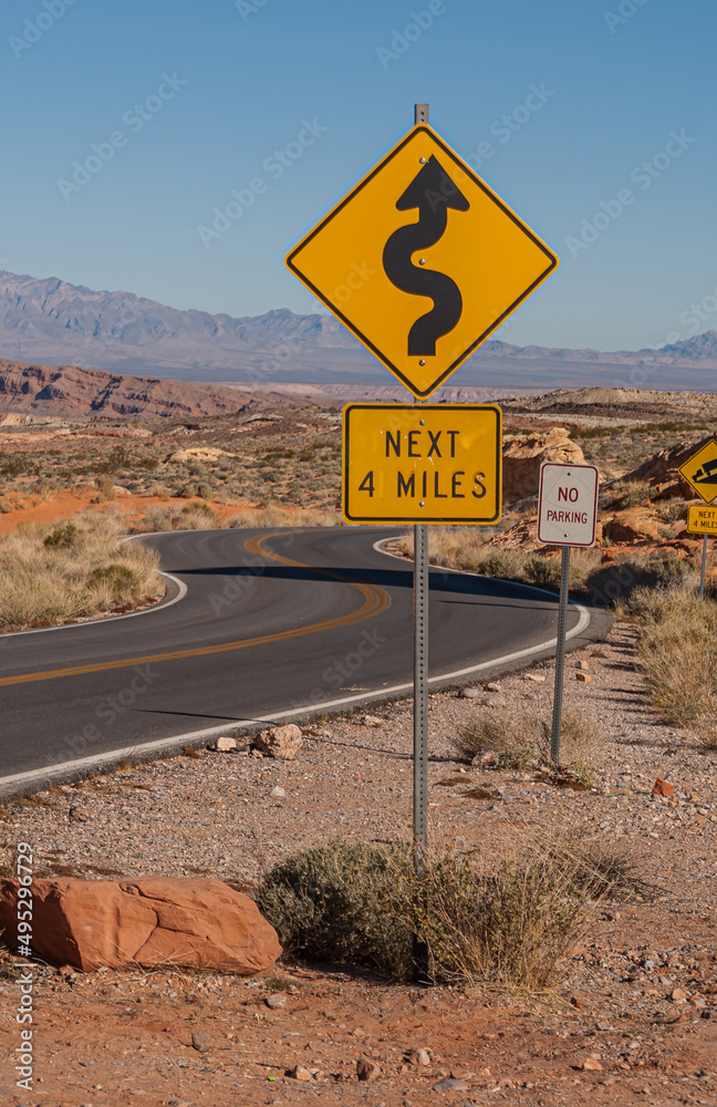 Overton, Nevada, USA - December 11, 2010: Valley of Fire. Black asphalt road meanders through wide brown mountain desert landscape. Portrait of yellow sharp turns road sign up front.