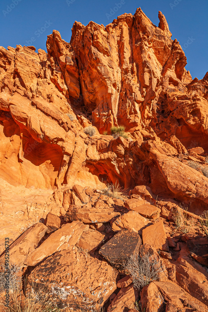 Overton, Nevada, USA - December 11, 2010: Valley of Fire. Group of sharp pinned red rocks close together behind heap of rocks with dry bushes on top under blue sky.