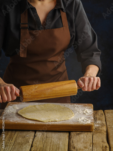 The chef rolls out the dough with a rolling pin on a wooden cutting board on a dark blue background. Close-up. Cooking pizza, pasta. Recipe concept. Restaurant, hotel, home cooking, pizzeria.