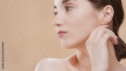 Dark-haired attractive young Caucasian beauty woman turns her head aside and touches jawline on beige background | Face care commercial concept