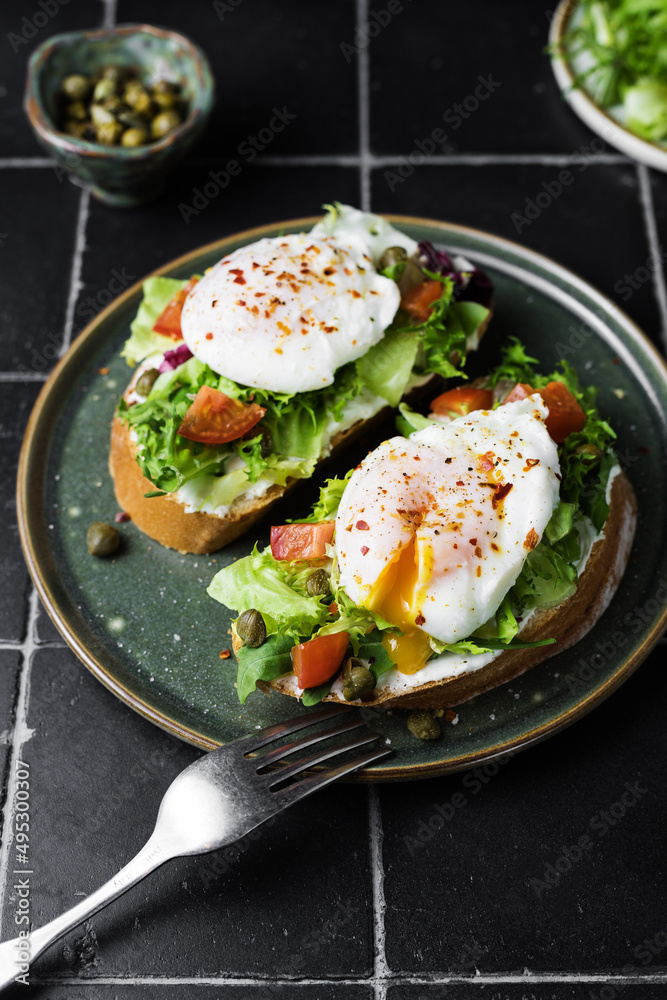 Toast with poached egg, lettuce, tomato and capers on a dark background, close up. Soft, selective focus Toast with poached egg, lettuce, tomato and capers on a dark background, close up. Soft, selective focus