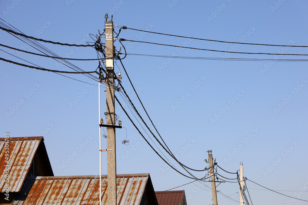 Powerline posts with electrical wires and capacitors above old roofs on blue sky background. Electricity transmission line, power supply in village