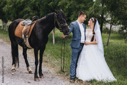 A young groom in a suit and a beautiful bride in a white long dress hug in a village outdoors, walking with a black horse along a country road. Wedding photography, portrait. © shchus