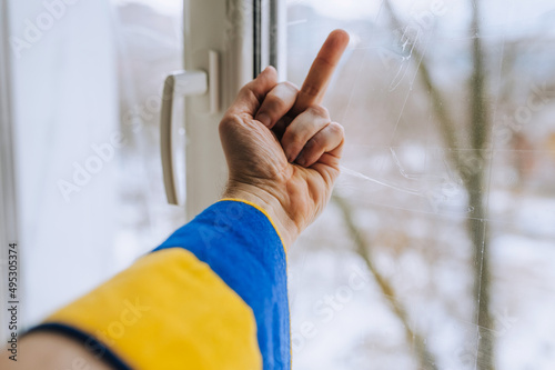 A Ukrainian man, being at home near a window in Ukraine under shelling in the war, shows an obscene gesture, the middle finger to the Russian occupiers. Concept, conflict.