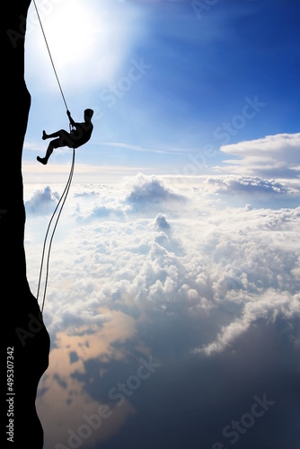Silhouette of young man abseiling down from a cliff high above clouds and sea, sun, beautiful colorful sky and clouds behind. Climber rappelling from a rock.