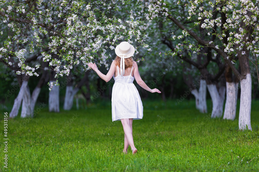 Spring landscape of apple covered with flower and green leaves. A woman in a hat, white sundress stands on the lawn. Beautiful, lush, flowering apple tree is dotted with flowers. Natural landscape.