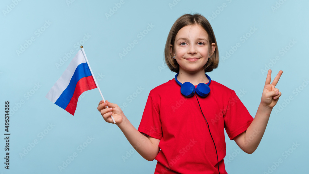 Nice girl 9-11 y.o. wearing basic red t-shirt and blue headphones on light blue background in studio showing winner gesture and peace sign. Child holding flag of Russia. Studying in Russia.