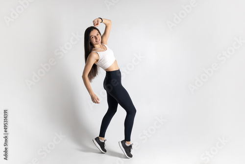 Full length view of smiling young woman posing and jumping on white background. Attractive brunette woman in fashionable sportswear exercising. Healthy lifestyle, sport concept