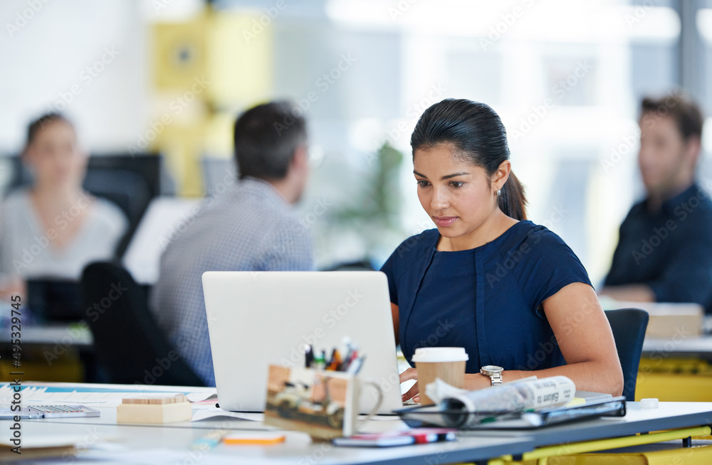 Immersed in her work. Shot of a designer sitting at her desk working on a laptop with colleagues in the background. Immersed in her work. Shot of a designer sitting at her desk working on a laptop with colleagues in the background.