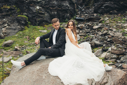 Portrait of a couple taking a photo shoot on their wedding day sitting on a stone back to back on the background of a rocky mountain hill. Beautiful portrait photo