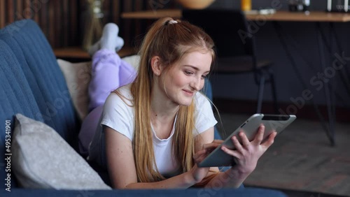 Caucasian good looking woman using tablet device and thinking while lying on the blue sofa at home. Beautiful girl lying on the couch and using digital tablet in living room. Female using gadget.